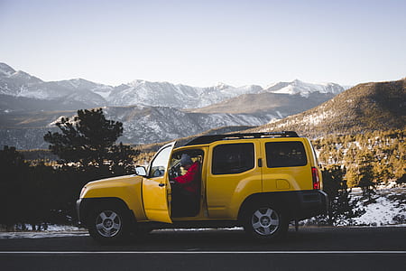 man wears red shirt in yellow SUV near mountain under cloudy sky thumbnail