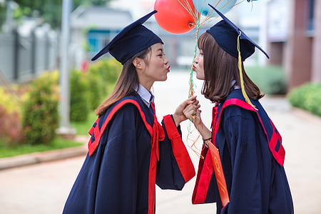 two women wearing black-and-red academic robe while facing each other while holding red and blue balloons thumbnail