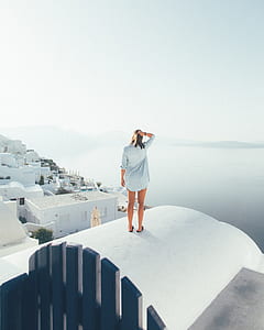 woman wearing blue long-sleeved shirt standing on white concrete building facing vast body of water thumbnail