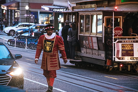 Man Wearing Red and Yellow Dress Walking on Concrete Road Near White and Red Cable Train thumbnail