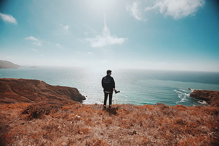 man standing on cliff facing body of water during daytime thumbnail