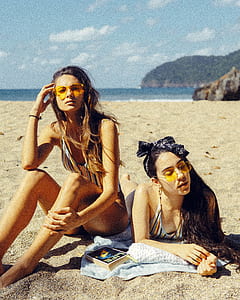 woman sitting on sand beside woman reclining near beach thumbnail