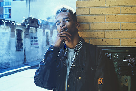 man wearing black jacket smoking behind brick wall in street side during daytime thumbnail