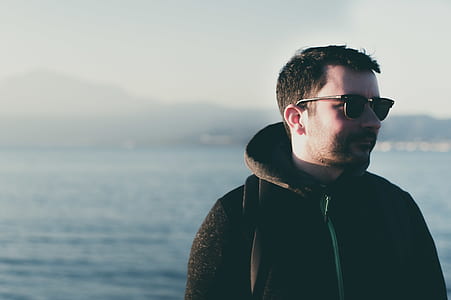 man wearing black sunglasses and hoodie looking sideways near body of water thumbnail