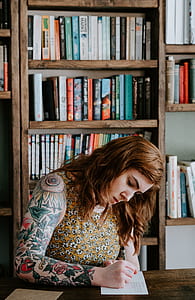 woman writing with green and red flower arm tattoo near bookcases thumbnail