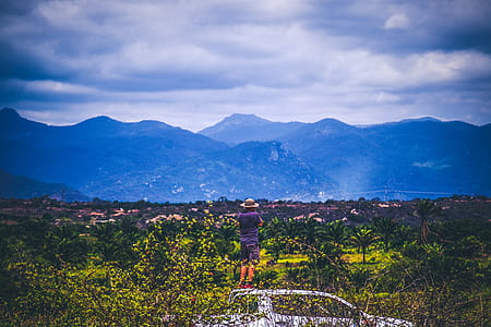 Person Wearing Sun Hat Looking At Mountains Under Cloudy Sky thumbnail