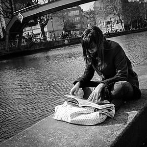 grayscale photography of woman reading book while sitting near body of water thumbnail