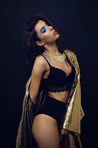 woman wearing black bralette and panty and gold-colored sequined coat standing in front of black wall thumbnail