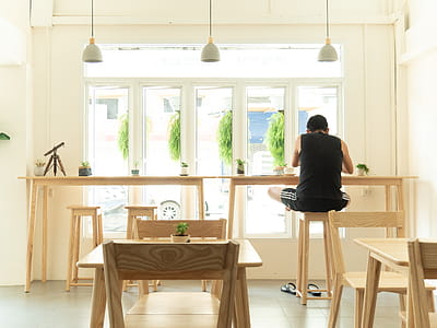 Man in Black Sleeveless Shirt Sitting on Wooden High Chair Beside Wooden Tables Near Window thumbnail