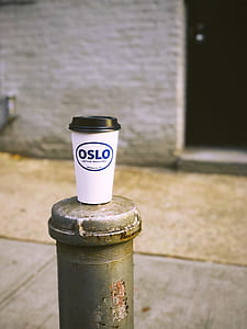tilt-shift lens photography of white and blue Oslo disposable coffee cup on top of gray metal pipe at side of a sidewalk during daytime thumbnail