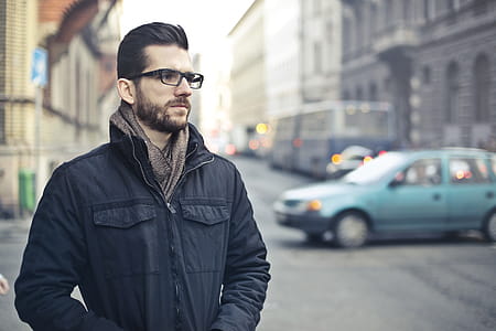 Young Bearded Man In Black Zip-Up Jacket and Spectacles, Thinking While Standing On The Street thumbnail