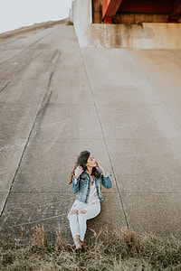 woman in blue washed denim jacket, white shirt, and distressed blue jeans leaning in concrete wall thumbnail