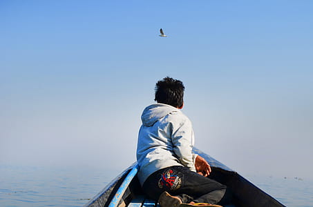 Man in Grey Hoodie and Black Pants Sitting in the Middle of Boat Looking at Bird in the Sky thumbnail