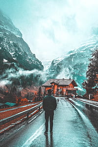 man standing on road facing house and mountains thumbnail