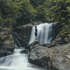 man in blue top standing on rock beside of water falls during daytime thumbnail
