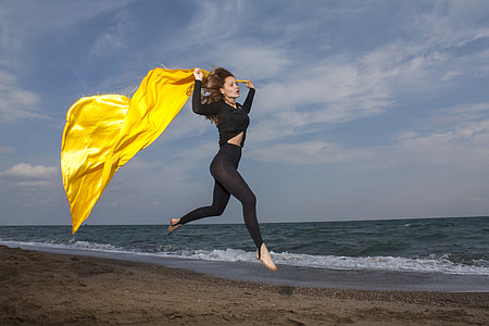 woman in black long sleeve tops and black leggings running through seashore holding yellow textile during daytime thumbnail