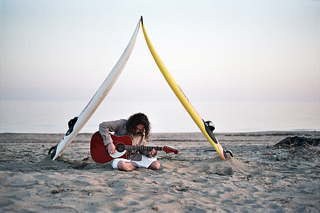 man playing red acoustic guitar with two white and yellow surfboard above his head by the beach thumbnail