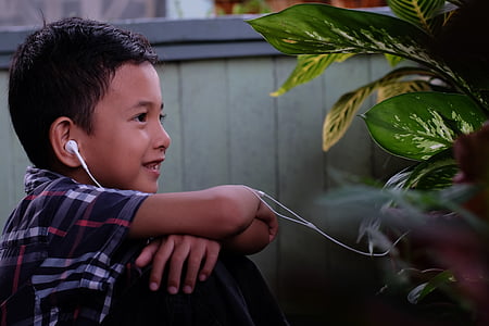 boy in black, red, and white plaid collared shirt and black bottoms outfit with white earphones on his ear while smiling at daytime thumbnail