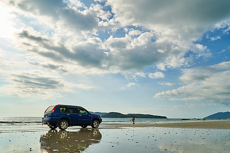 blue SUV near person standing on seashore in distant of mountains thumbnail