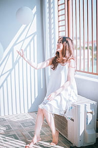 woman wearing white sleeveless dress playing white ball while sitting on outdoor AC thumbnail