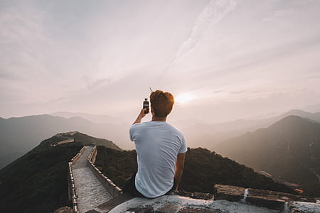 man in white T-shirt sitting on edge of wall while holding black Android smartphone thumbnail