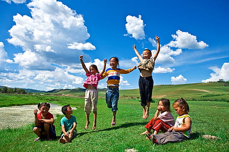 four children sitting on green grass and three children jumping taken under white clouds during daytime thumbnail
