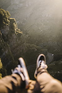 person wearing black and white sneakers sitting on cliff fronting curve road in between mountains thumbnail