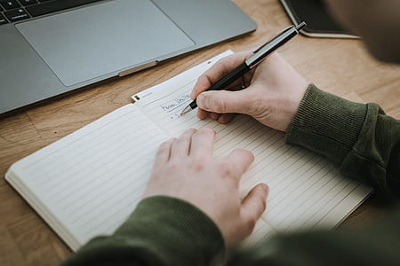 person holding black click pen writing on book beside gray laptop computer thumbnail