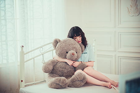 woman in white top hugging brown bear plush toy on bed thumbnail