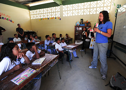 man in blue shirt and gray pants standing in front of children inside room with whiteboard thumbnail