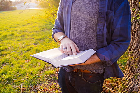person standing near tree holding book while reading during daytime thumbnail