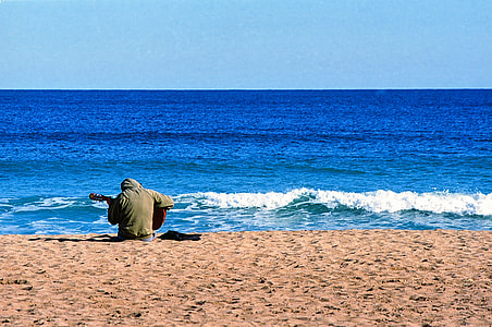 man holding guitar sitting on sand near body of water thumbnail
