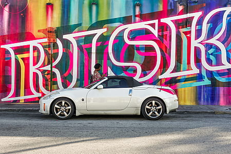 woman standing behind white Nissan 350Z convertible coupe near graffiti filled wall thumbnail