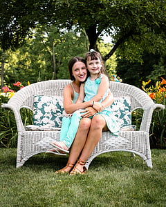 child in teal sleeveless shirt and pants sitting on woman lap in teal sleeveless dress sitting on white wicker sofa thumbnail