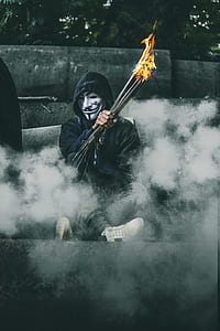 man in fawkes mask holding burning rods surrounded by smoke thumbnail
