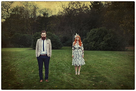 red-haired woman standing wearing white dress beside bearded man near trees thumbnail
