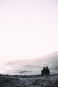 two persons sitting near seashore during foggy day thumbnail