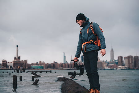 man standing on black tree trunk capturing video of seawaves through video recorder during daytime thumbnail