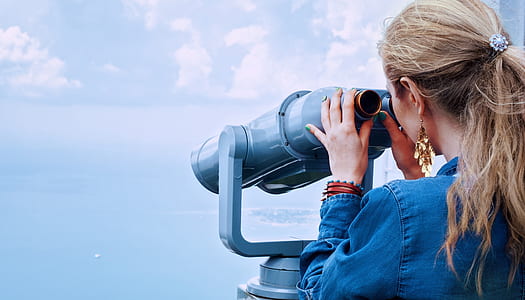 man wearing blue denim long-sleeved top holding black tower telescope in front of body of water thumbnail