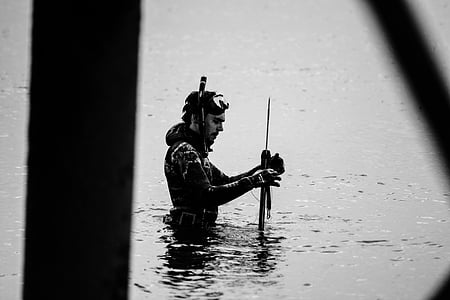 grayscale photo of man holding spear in body of water thumbnail