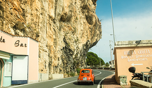 red vehicle on gray concrete road between brown concrete building and brown rock formation during daytime thumbnail