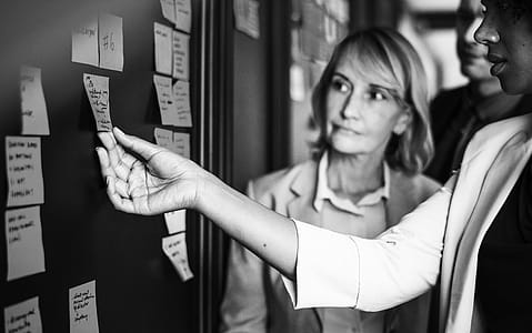 Grayscale Photography of Woman Looking at Woman Picking Up Sticky Note on Board thumbnail