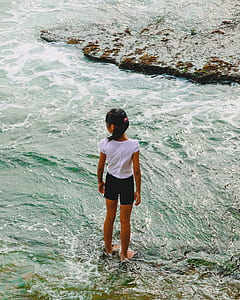 girl in white shirt and black shorts standing in ankle deep water near rock at daytime thumbnail
