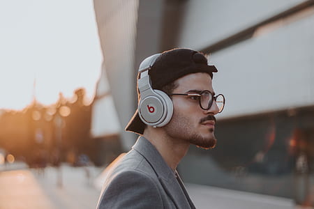 tilt shift lens photography of man wearing black frames round eyeglasses, black snapback cap and gray sport coat looking right with distance at gray building thumbnail