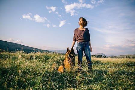 woman wearing scoop-neck long-sleeved shirt standing on green grass field beside tan and black German shepherd during daytime thumbnail