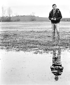 Man in Jacket Standing Near Body of Water Holding Camera in Grayscale Photography thumbnail