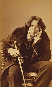 man wearing black suit and black coat holding cane sitting on chair thumbnail