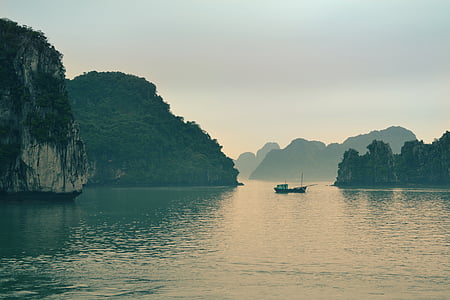 boat on calm body of water near rock formation covered by trees under white sky during daytime thumbnail