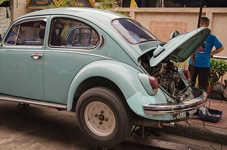 man standing on sidewalk in front of blue Volkswagen Beetle coupe parked on side of the road with open hood exposing engine thumbnail