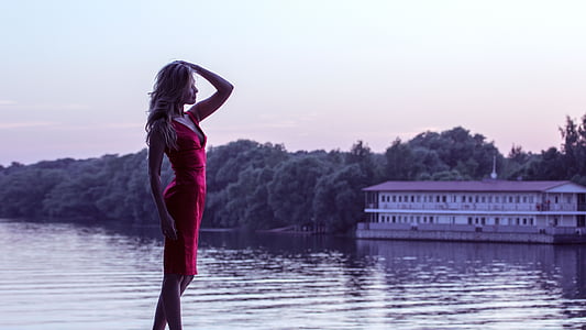 woman wearing red mini dress standing beside body of water during golden hour thumbnail